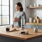 whb-superb AeroPress Premium Kaffeepresse aus Glas und Edelstahl auf einer Holzküche mit Frau im Hintergrund
