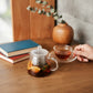 whb-superb Glasteekanne mit Teesieb und Früchtetee auf einem Holztisch neben Büchern und einer Teetasse