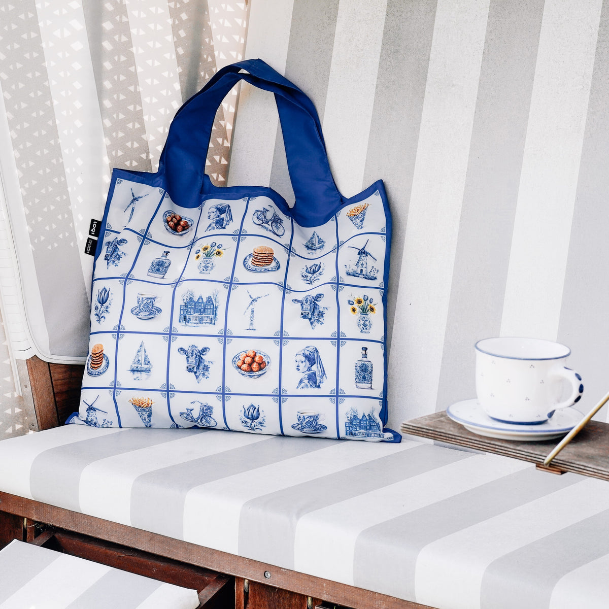 whb-superb LOQI Tasche mit blauem Delfter Kachelmuster aus recyceltem Polyester auf einer gestreiften Bank