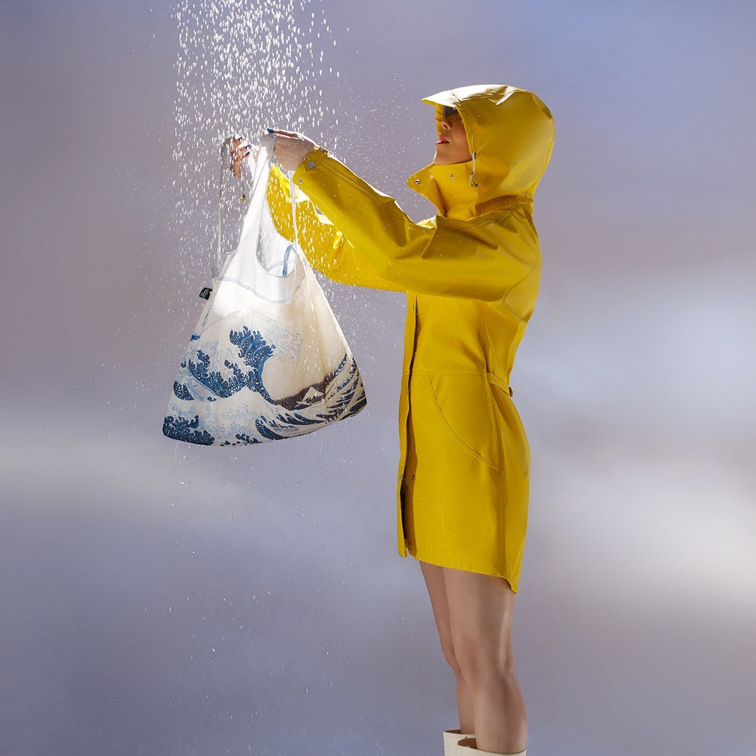 whb-superb LOQI Einkaufstasche aus recyceltem Taft mit Fuji-Design, gehalten von einer Frau im gelben Regenmantel