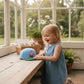 Ein kleines Mädchen lächelt neben einer hellblauen whb-superb Peppa Pig Snapback Cap mit grünem Schirm auf einem Holztisch.